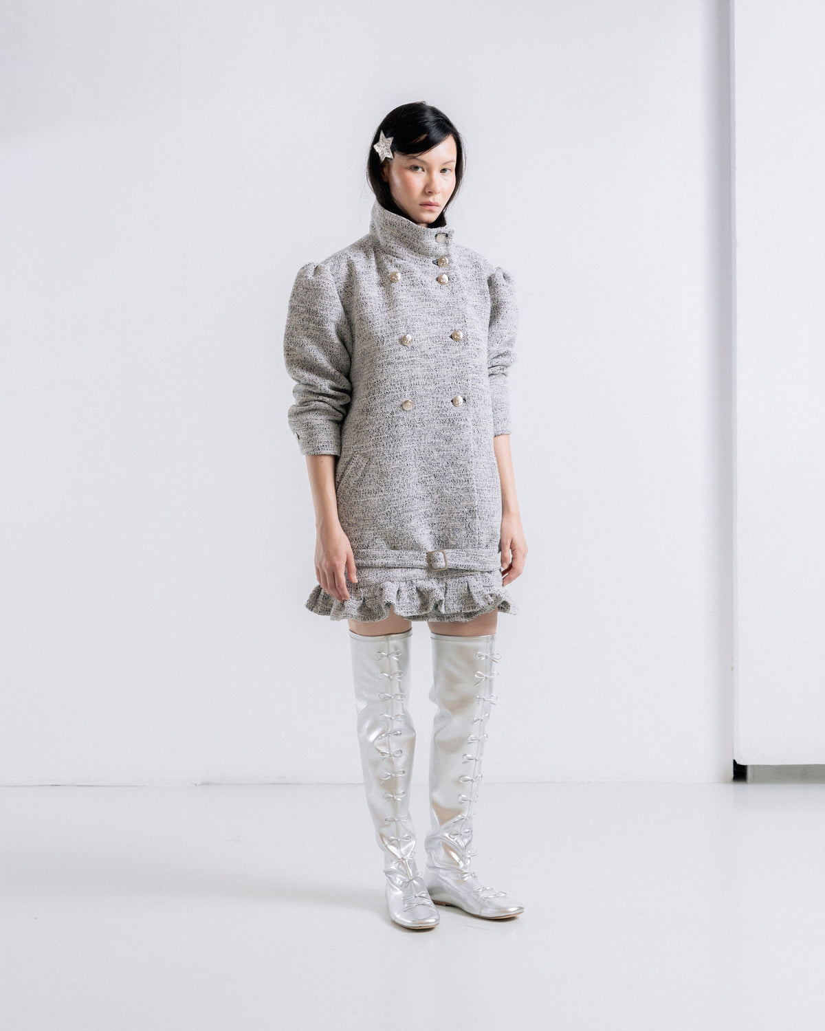 A model stands in a minimalist white studio setting wearing a gray tweed double-breasted mini dress with a high mock neck and ruffled hem, paired with shiny silver thigh-high boots.