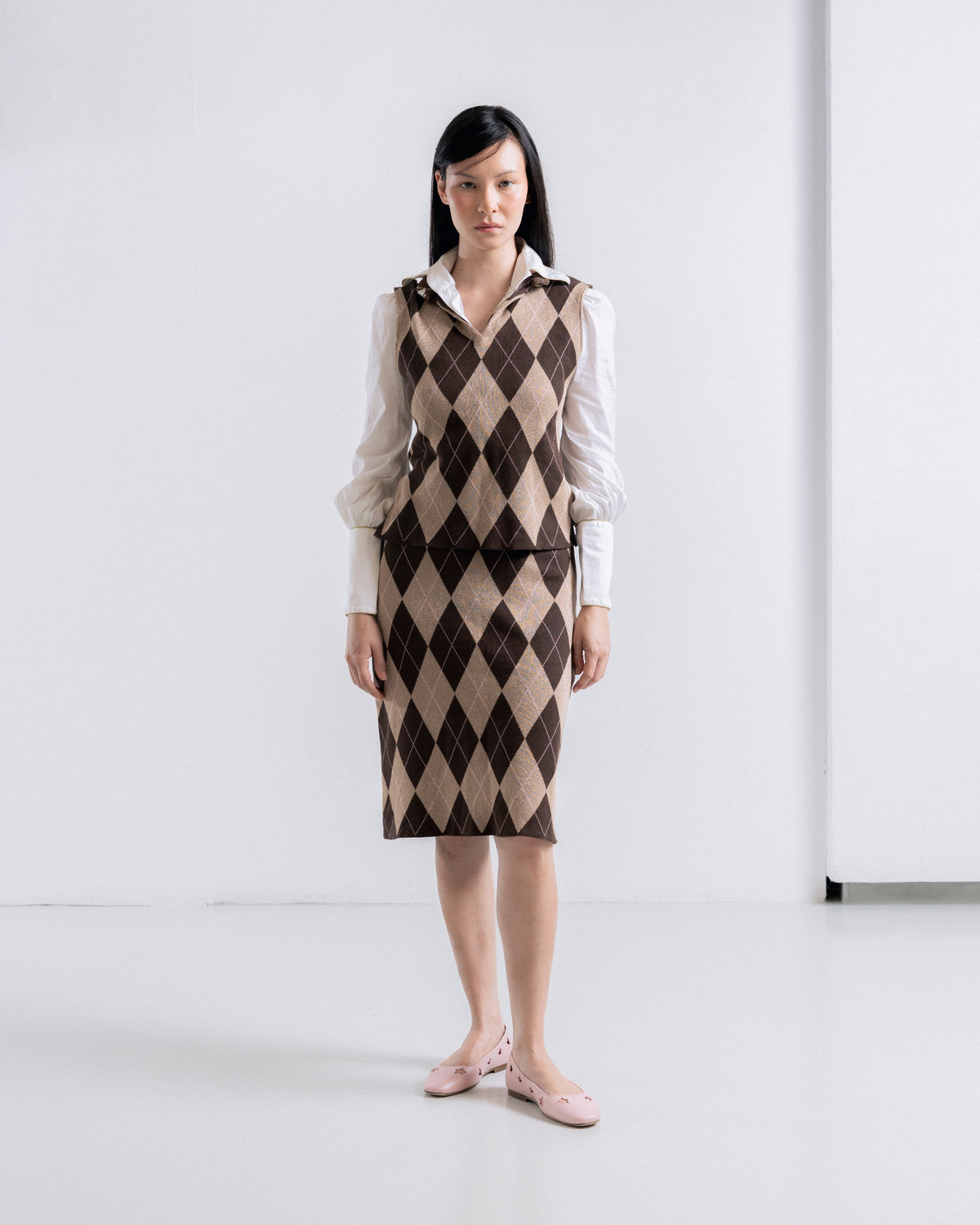 A woman stands in a studio wearing a sleeveless collared dress with an argyle pattern in brown and beige over a white blouse, paired with pale pink flats.