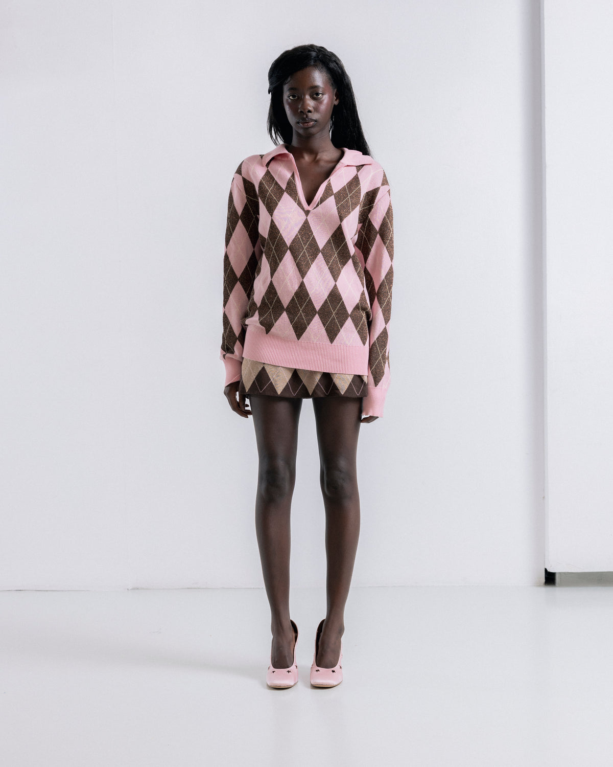 A model stands in a studio wearing a pink and brown argyle knit sweater with a matching mini skirt and pink heels.