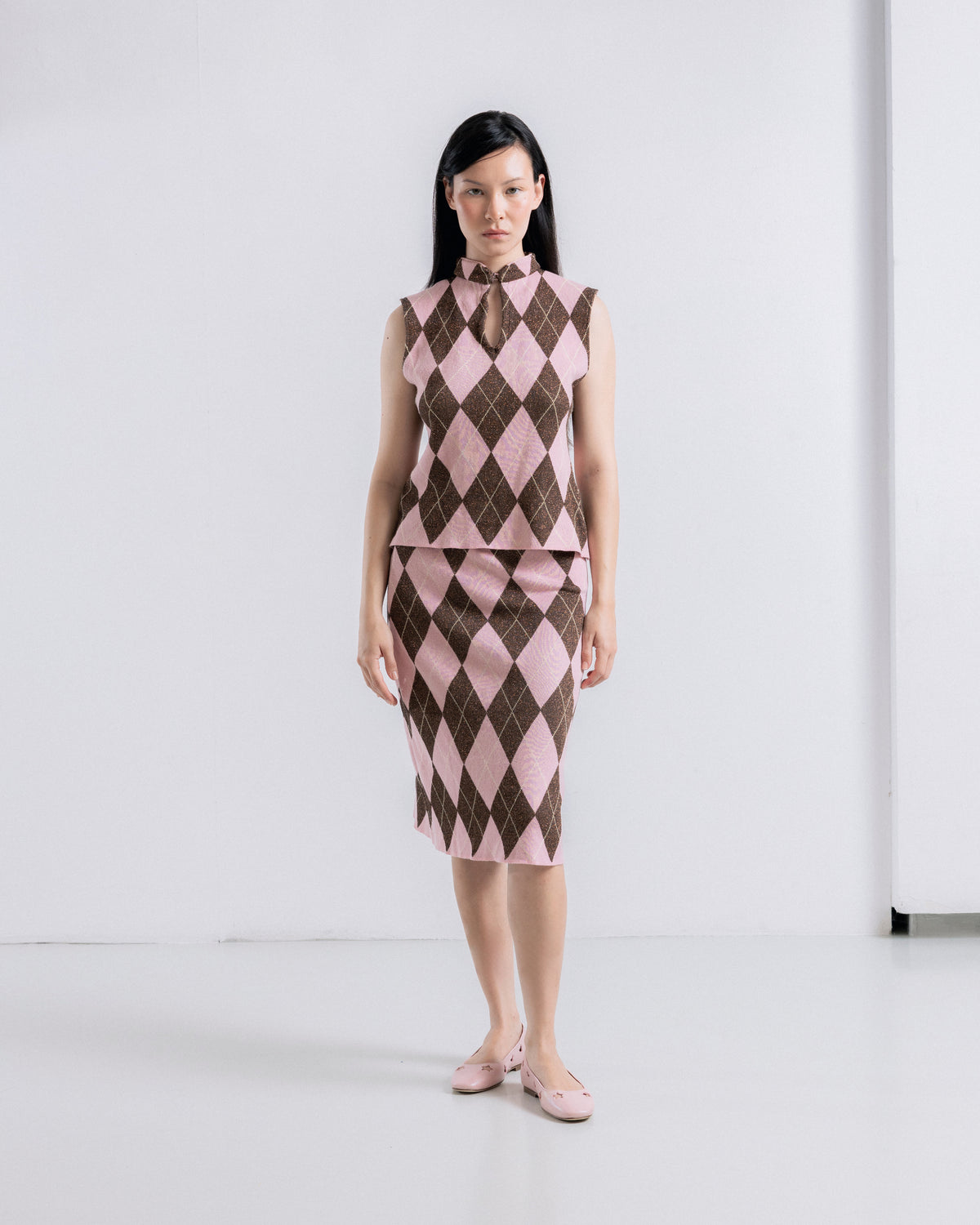 A model stands in a studio wearing a sleeveless high-neck dress with a brown and pink argyle pattern, paired with matching pink shoes.