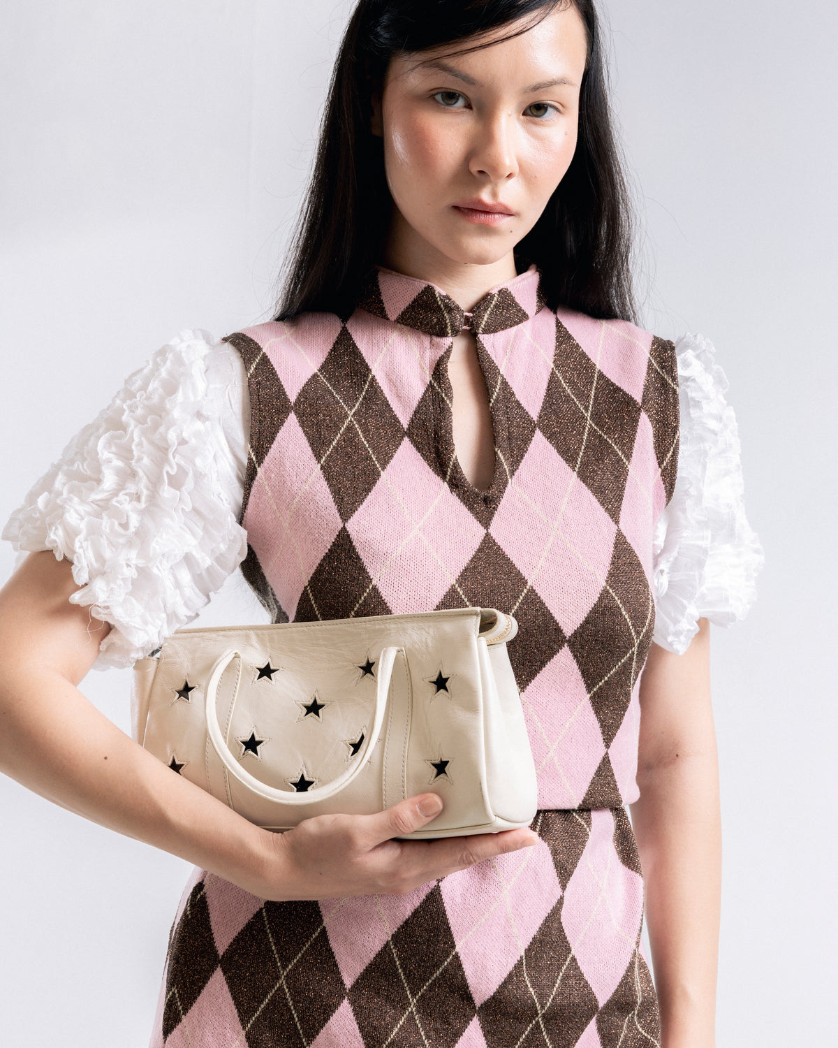 A woman wearing a pink and brown argyle sweater vest with white ruffled short sleeves holds a cream-colored handbag decorated with black stars, set against a plain light gray background.