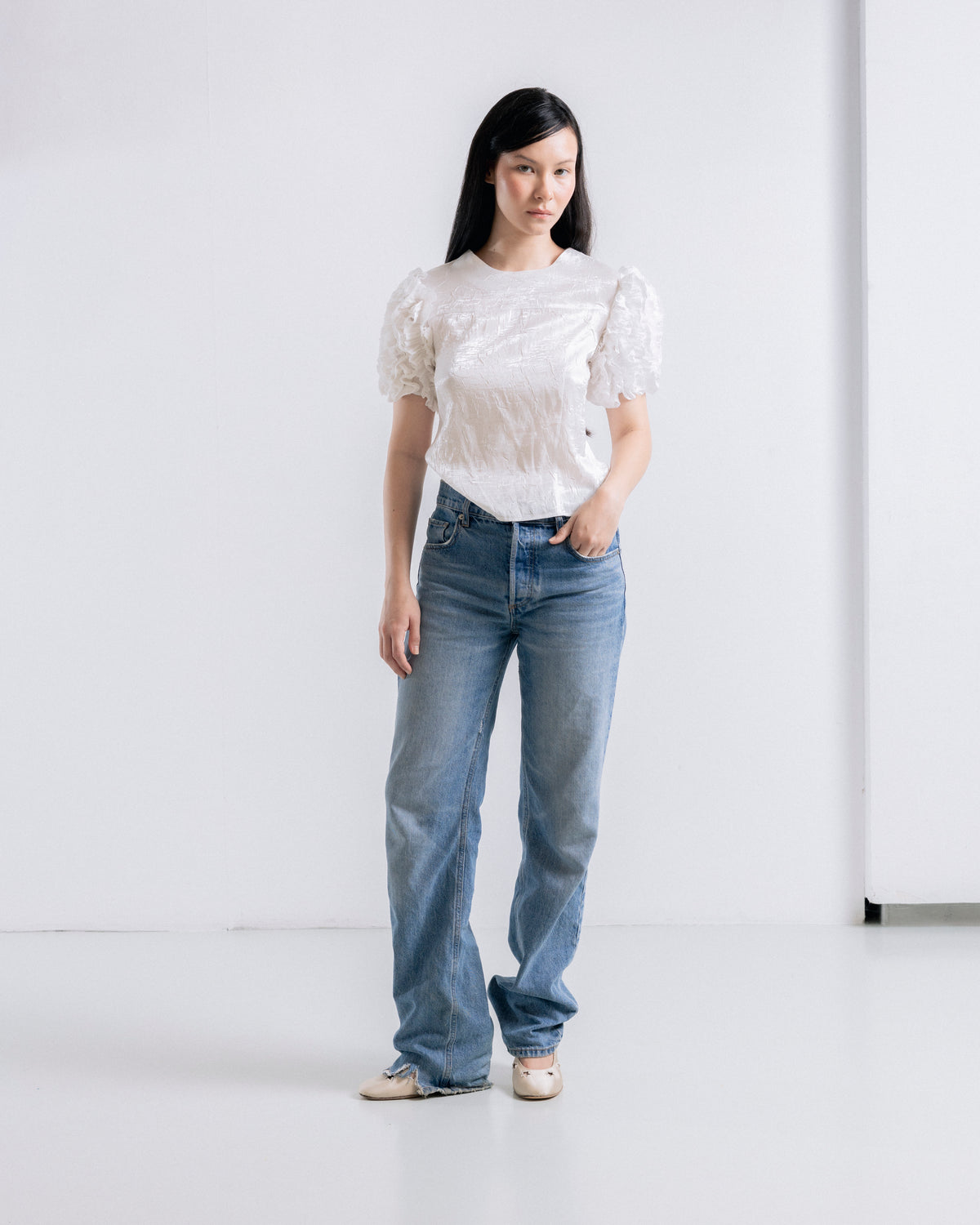 A woman stands in a bright white studio wearing a white short-sleeve lace top, blue jeans, and beige shoes.