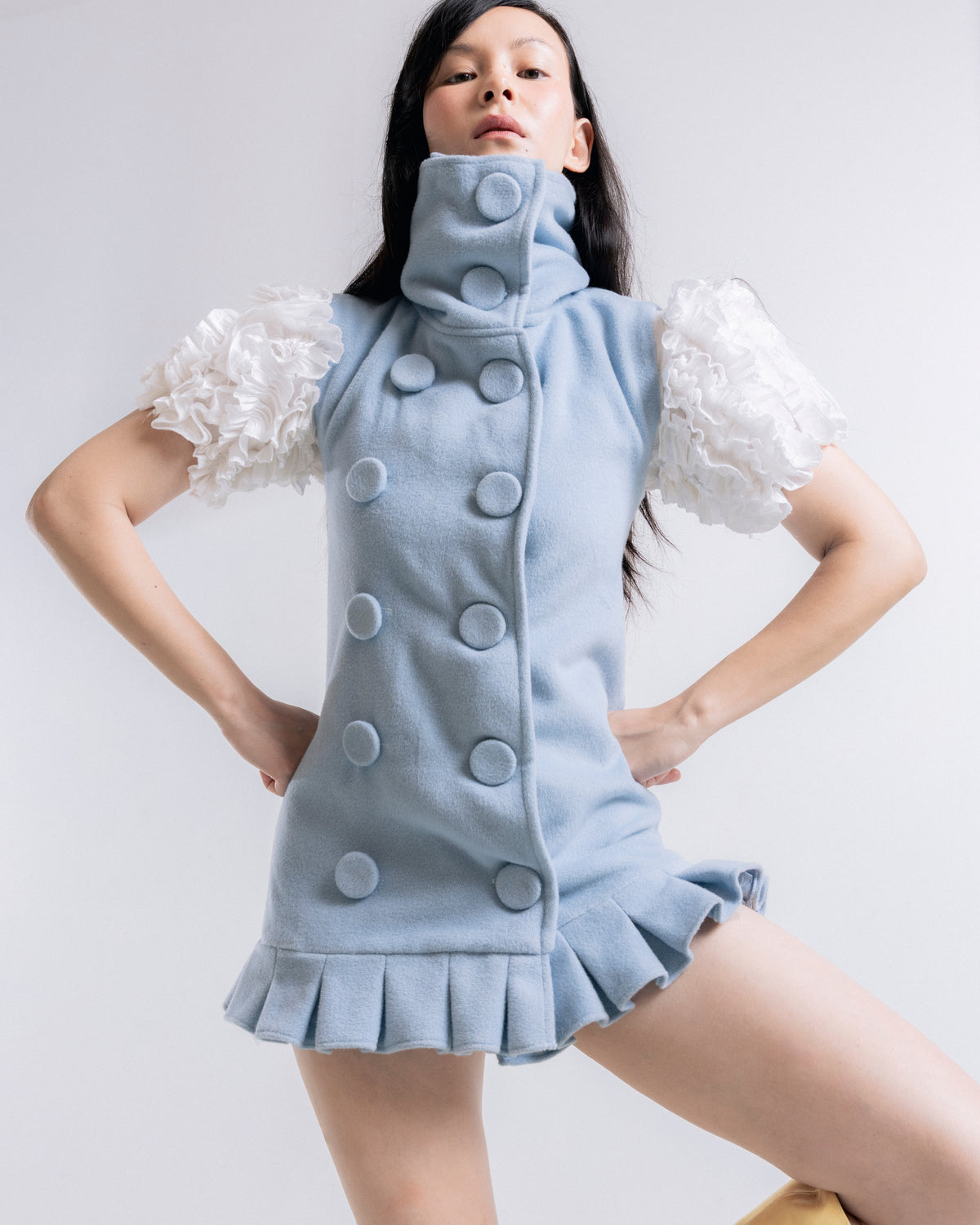 A model wearing a light blue high-neck mini dress with oversized decorative buttons, ruffled white puff sleeves, and a ruffled hem, posed against a light gray studio background.
