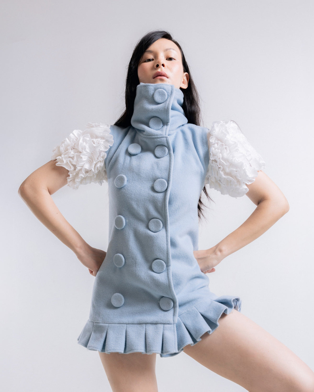A model wearing a light blue sleeveless mini dress with a high mock neck, large decorative buttons, ruffled white puff sleeves, and a ruffled hem, posed against a light gray studio background.