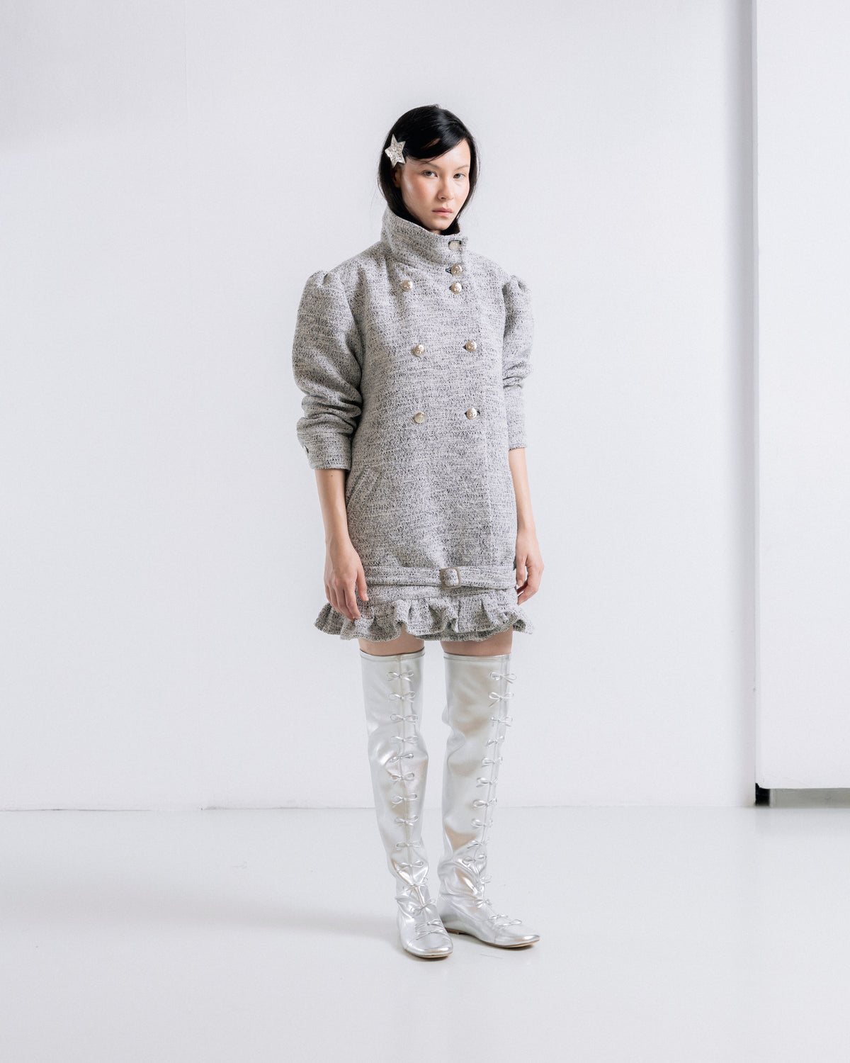 A model stands in a minimalist white studio setting wearing a gray tweed double-breasted mini dress with a high mock neck and ruffled hem, paired with shiny silver thigh-high boots.